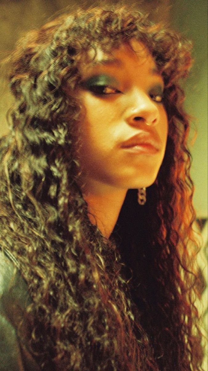 a closeup portrait of a young woman with curly hair, wearing green eyeshadow and a chain earring. she is looking off to the side and her expression is serious.