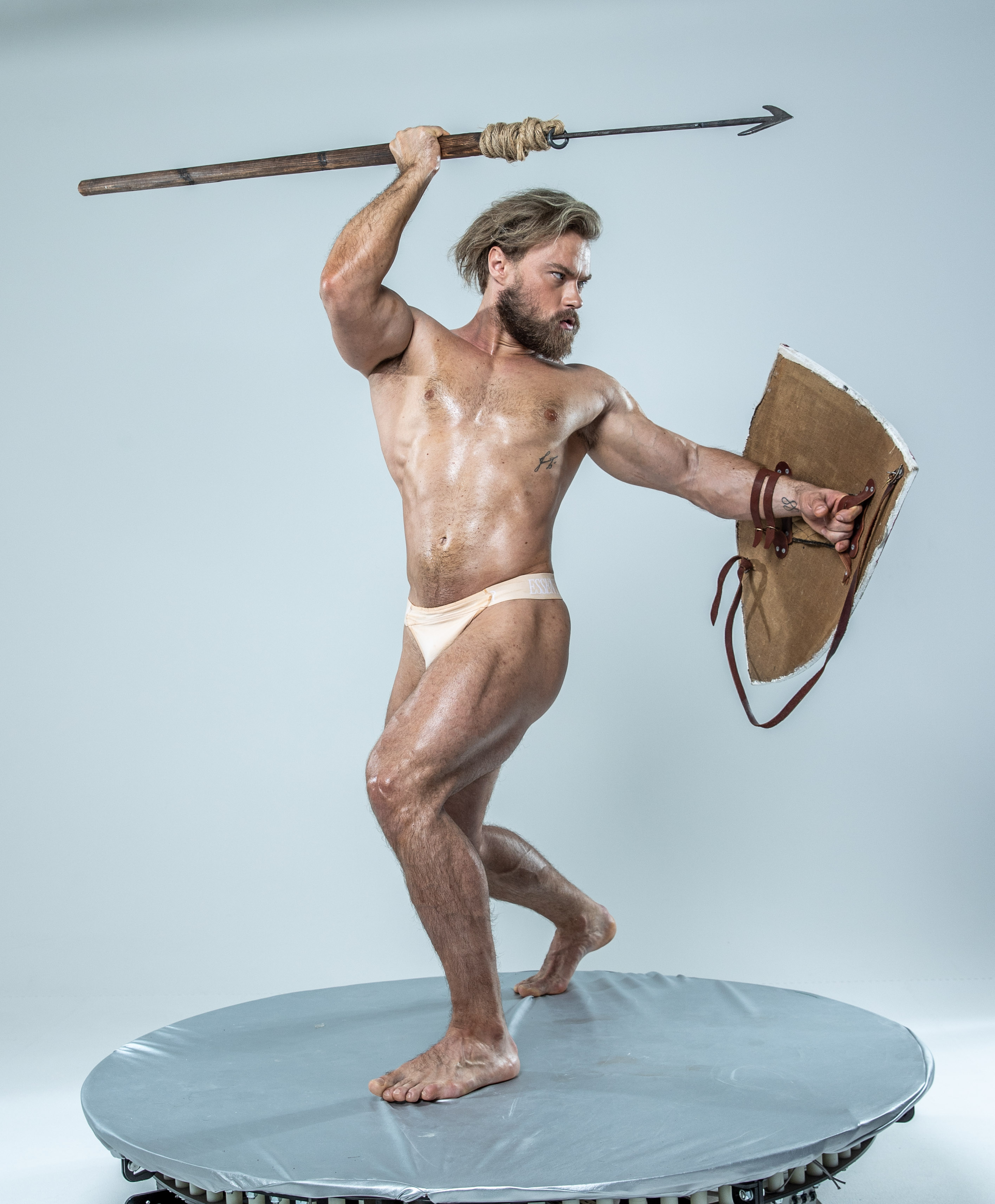 a muscular man, his body glistening with sweat, stands poised for battle on a circular platform. he brandishes a spear in his right hand, held high above his head, and a round shield in his left. his stance exudes strength and power, the embodiment of an ancient warrior. the studio setting, with its plain white background, emphasizes the man's physique and the intensity of his pose.