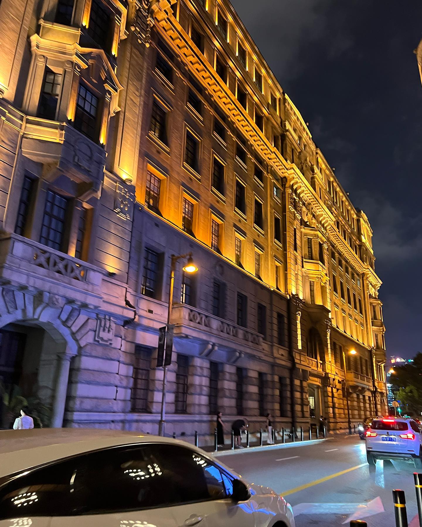 the image captures a tall, grand building illuminated by warm, golden lights against a dark night sky.  it exudes a sense of history and grandeur. the street in front is moderately busy with cars and a few pedestrians. the perspective is from street level, giving a sense of the building's scale.