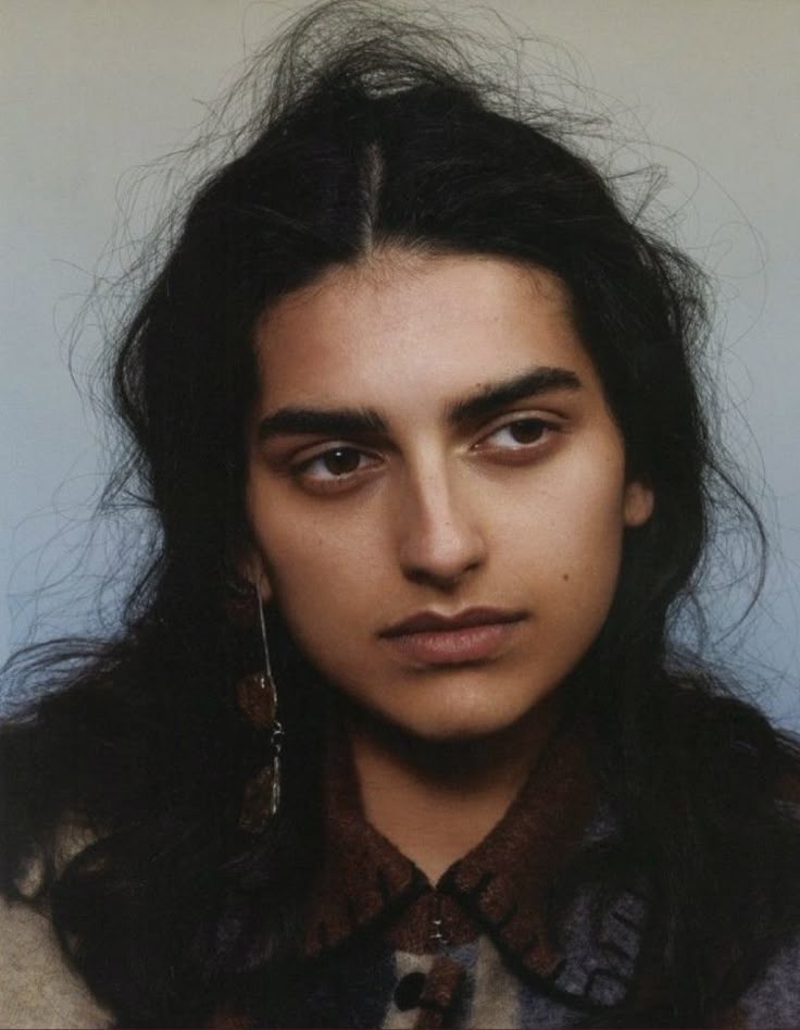 a portrait of a woman with dark hair parted in the center, thick eyebrows, and brown eyes. she is wearing long earrings and a patterned shirt, and has a serious expression on her face.
