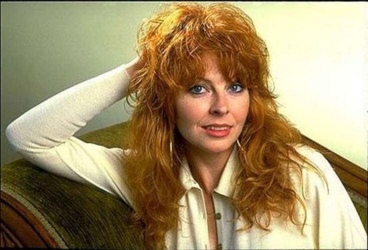 a woman with long, wavy red hair is looking at the camera and smiling. she is wearing a white shirt with a collar. the background is a light, neutral color.
