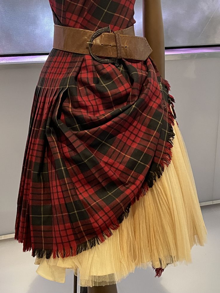 closeup shot of a mannequin wearing a plaid dress with a brown belt. the dress is made of red and black plaid fabric with pleats and fringe. the belt is brown and has a large buckle. the mannequin is standing in front of a lightcolored wall. the dress also has a tan, tulle underlayer.