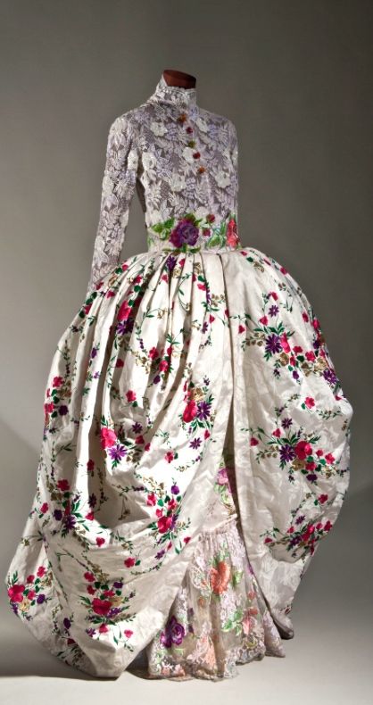 a photograph features a mannequin dressed in a vintagestyle gown, which is likely formal wear. the upper portion of the gown is made of lace with a high neckline and long sleeves, adorned with a few buttons down the front. a floral waistband defines the transition to the voluminous skirt. the skirt is predominantly white with a floral pattern consisting of red, pink, and purple flowers with green leaves. the lower part of the skirt has a ruffled lace trim. the overall style suggests a historical period dress.