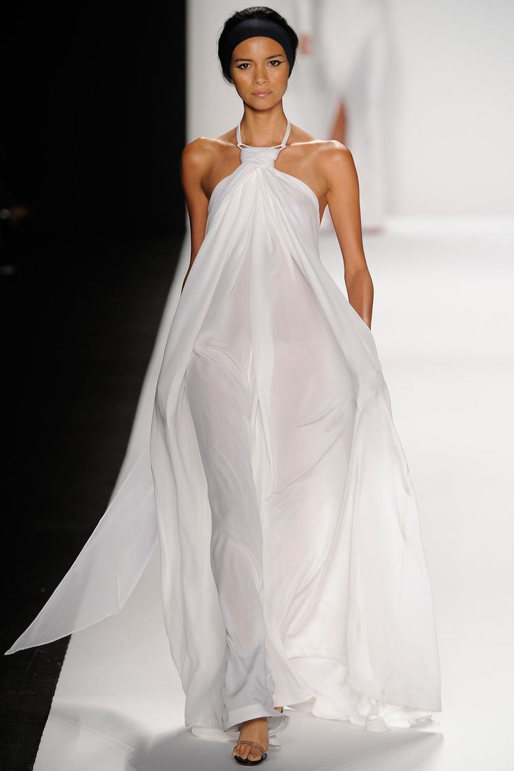 a fashion model walks the runway wearing a long, flowing white dress. she has a black headband in her hair. the dress is very sheer and drapes beautifully. she is posing for the camera with her legs slightly crossed and her arms at her sides. the model is wearing simple sandals.  the runway is white and the background is blurry.  she is wearing a very stylish and elegant outfit.