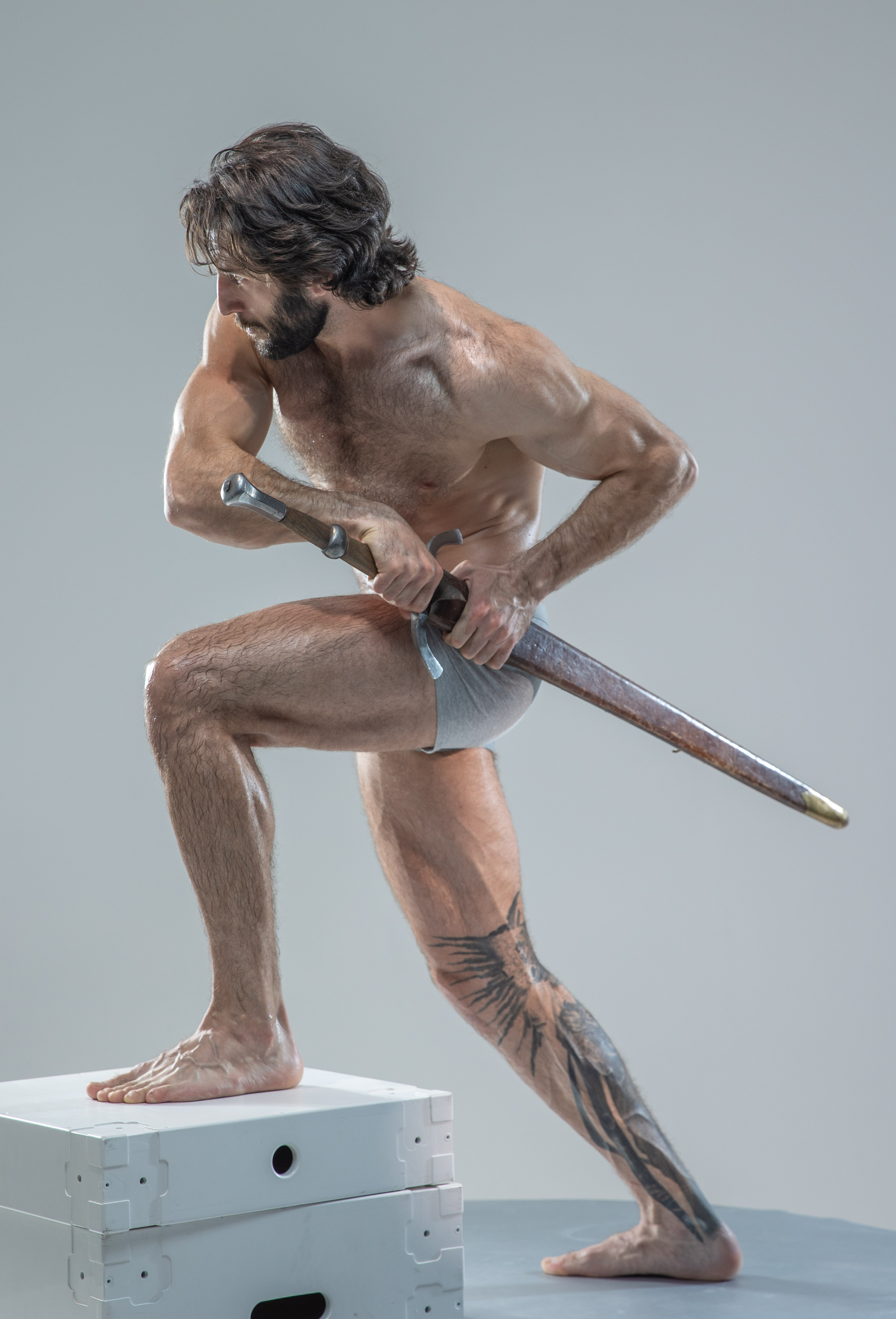 a muscular man with long hair and beard stands on a white platform in a classic warrior pose. he holds a sword with both hands, his body turned to the side, suggesting impending action. his physique is athletic and defined, with a tattoo visible on his lower leg. the background is a neutral gray, emphasizing the man and his powerful stance. the soft lighting enhances the definition of his muscles and creates a dramatic atmosphere.