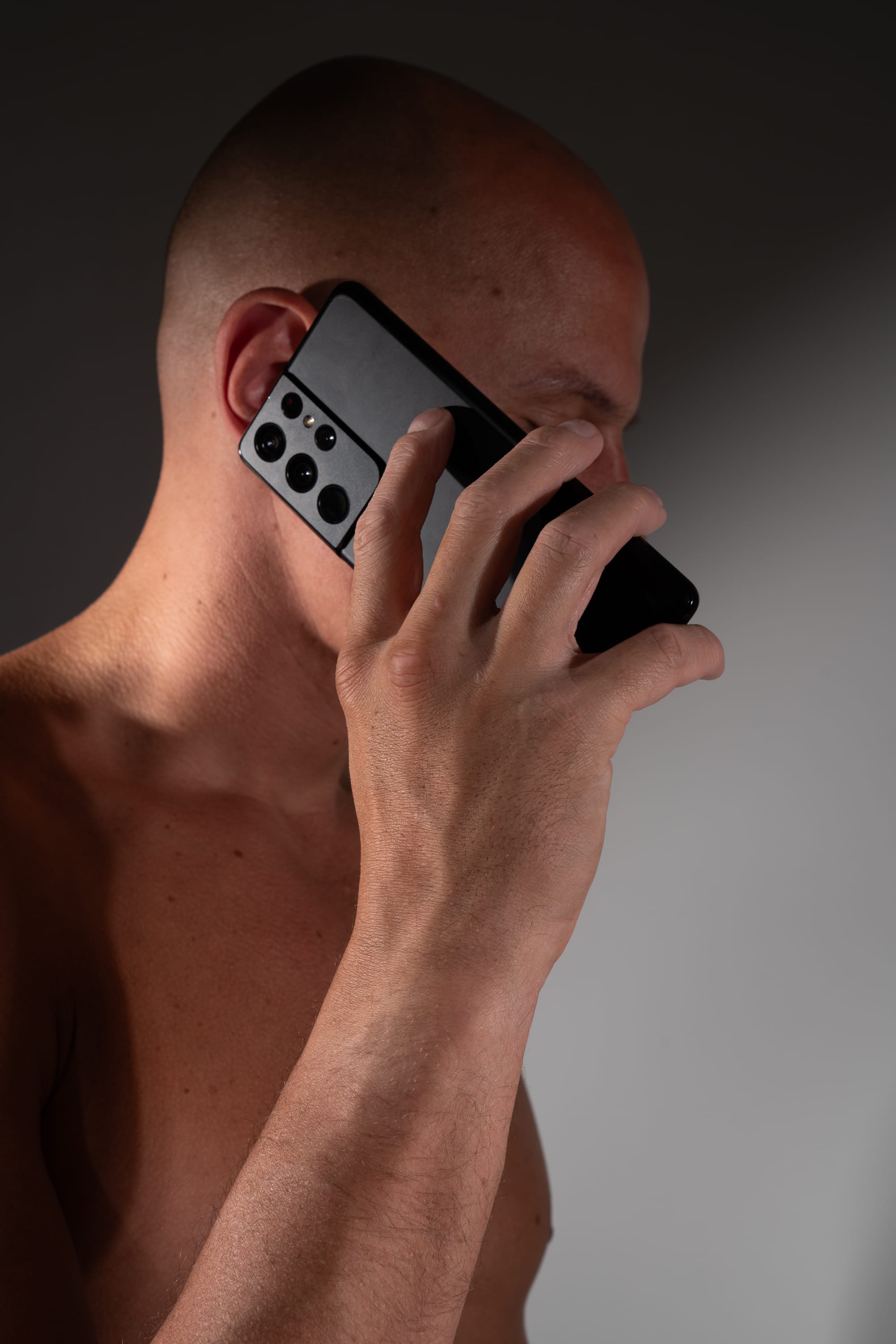 the image showcases a shirtless, bald man engaged in a phone conversation. he is positioned sideways, highlighting his upper body, and his expression is obscured as he listens intently to the call. the studio lighting creates a play of light and shadows across his body, adding depth to the image. the focus remains on the man's interaction with the smartphone, emphasizing the act of communication.