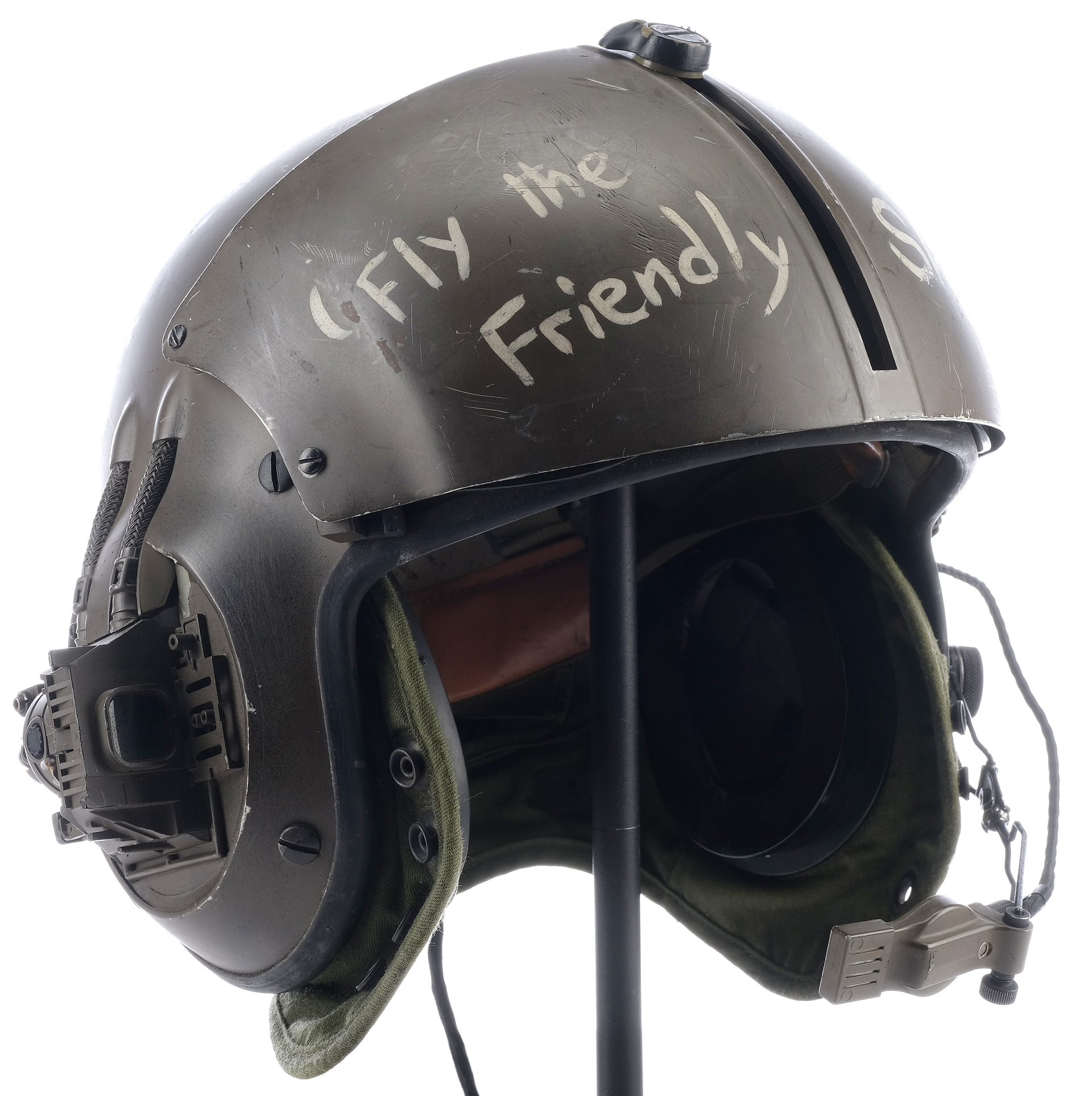 a closeup of a green military pilot's helmet, with the words "fly the friendly skies" written in white marker on the side. the helmet is equipped with a visor, goggles, headset, and microphone.