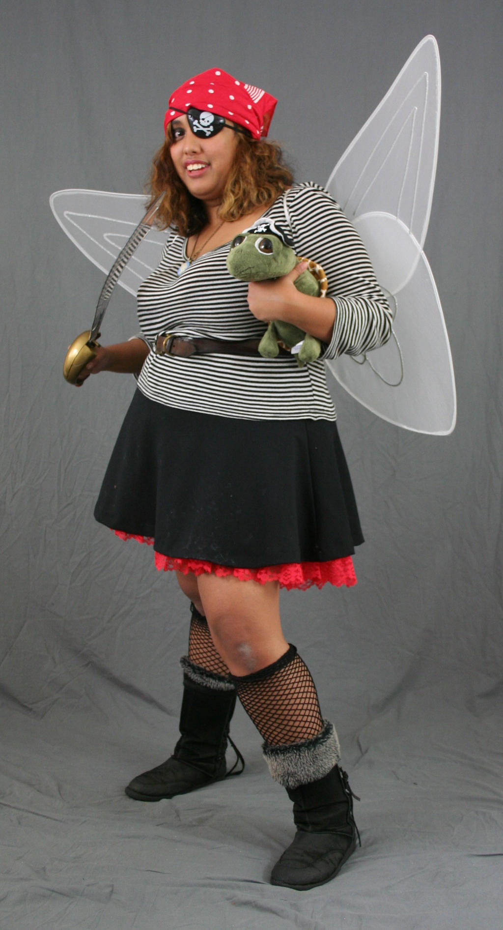 a full shot of a woman dressed in a pirate fairy costume. she is wearing a red bandana, an eyepatch, a striped shirt, a black skirt, and fishnet stockings with boots. she is holding a toy turtle and has large white fairy wings.  she is holding a sword.  the background is gray.
