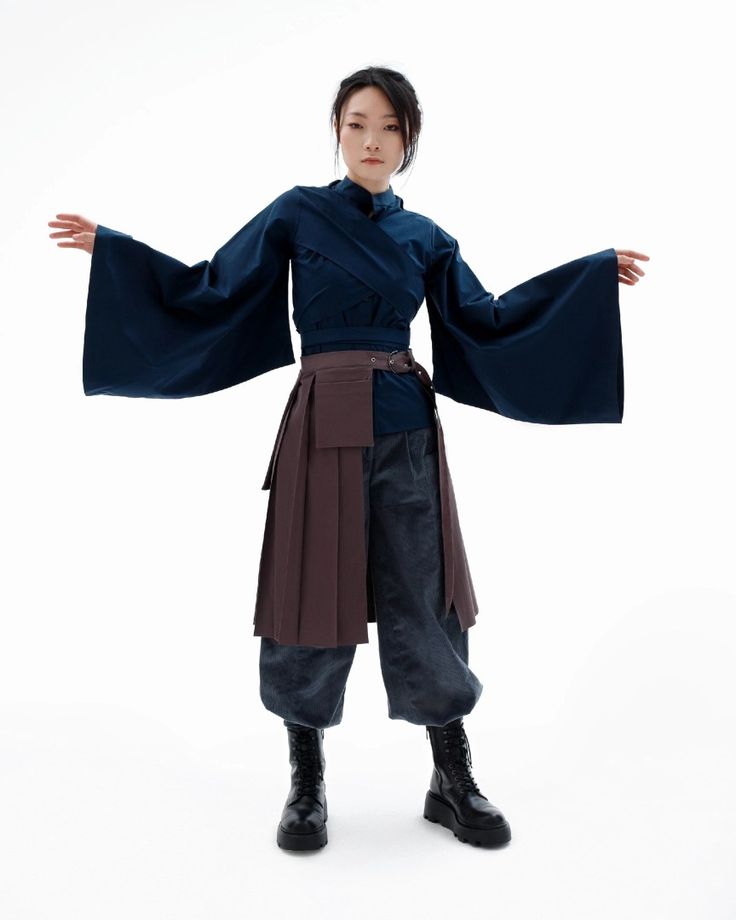 a woman is standing in a studio with a white background, she is wearing a blue, black, and brown outfit, and she is looking at the camera. she is wearing a blue top with wide sleeves and a brown skirt with a black belt. she is also wearing black boots.