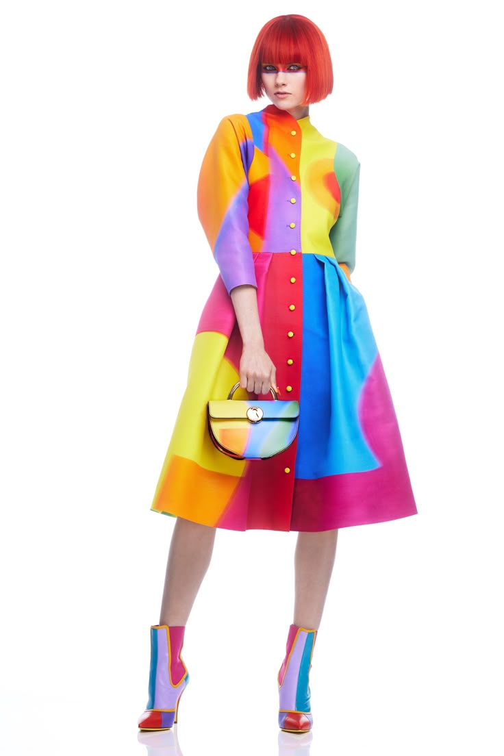 a woman is standing in a colorful dress and boots, holding a handbag, against a white background. she has short red hair. the dress has a pattern of geometric shapes and circles in various colors including orange, yellow, red, green, blue and pink. the handbag matches the colors of the dress. the boots are also colorful with vertical stripes.