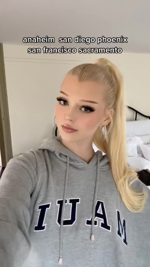 a woman with blonde hair and makeup is wearing a gray hoodie with "ijam" written on it. she has earrings and her hair is in a ponytail. the background shows a bed and the words "anaheim san diego phoenix san francisco sacramento" are written above her head.