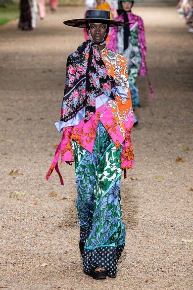 the image shows a fashion model walking down a gravel runway during an outdoor fashion show. the model is wearing a striking outfit composed of a wideleg pantsuit featuring a mix of vibrant floral and paisley prints in various shades of pink, green, blue, and orange. the outfit is styled with a large black hat, a long, flowing black and floral print scarf that is intricately draped around the neck and chest area.  other models can be seen in the background wearing similar, though less patterned, outfits. the overall aesthetic suggests a bohemian or eclectic highfashion style.