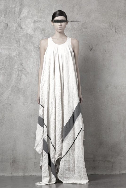 a full shot of a slender woman standing in front of a concrete wall, wearing a long, loosefitting white dress with grey and black accents. she has a blurred black mark across her eyes, resembling a blindfold.