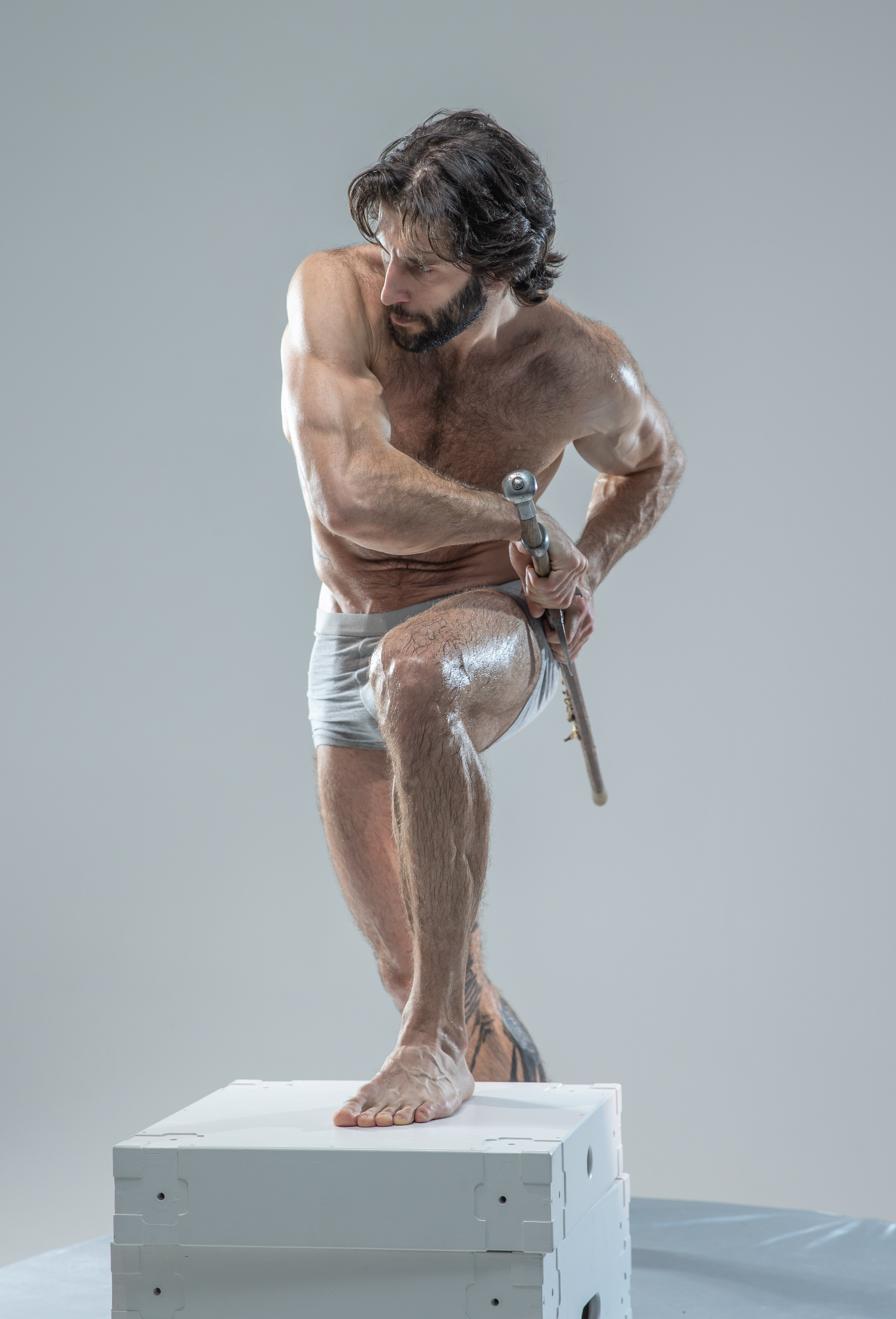 a muscular man with long dark hair and beard is posing on a white cube in a studio setting. he's wearing white underwear and is holding a sword in his hands. the background is a light gray.