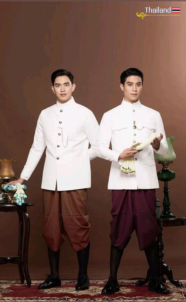 two men are standing in front of a brown wall. they are both wearing traditional thai clothing. they are both smiling and look very happy.