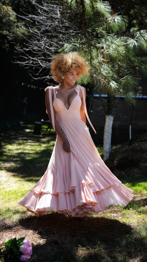 a young woman with voluminous curly blonde hair smiles gently as she stands in a sundappled garden. she is wearing a beautiful, light pink maxi dress with a tiered ruffled skirt and a vneckline. the dress has thin straps that tie at the shoulders.  the setting is lush and green, with tall trees and shadows creating a natural, idyllic backdrop.  a bouquet of pink flowers lies on the ground near her feet, adding to the overall serene and romantic ambiance of the photograph.  the image showcases the dress, her hair and the natural setting beautifully.