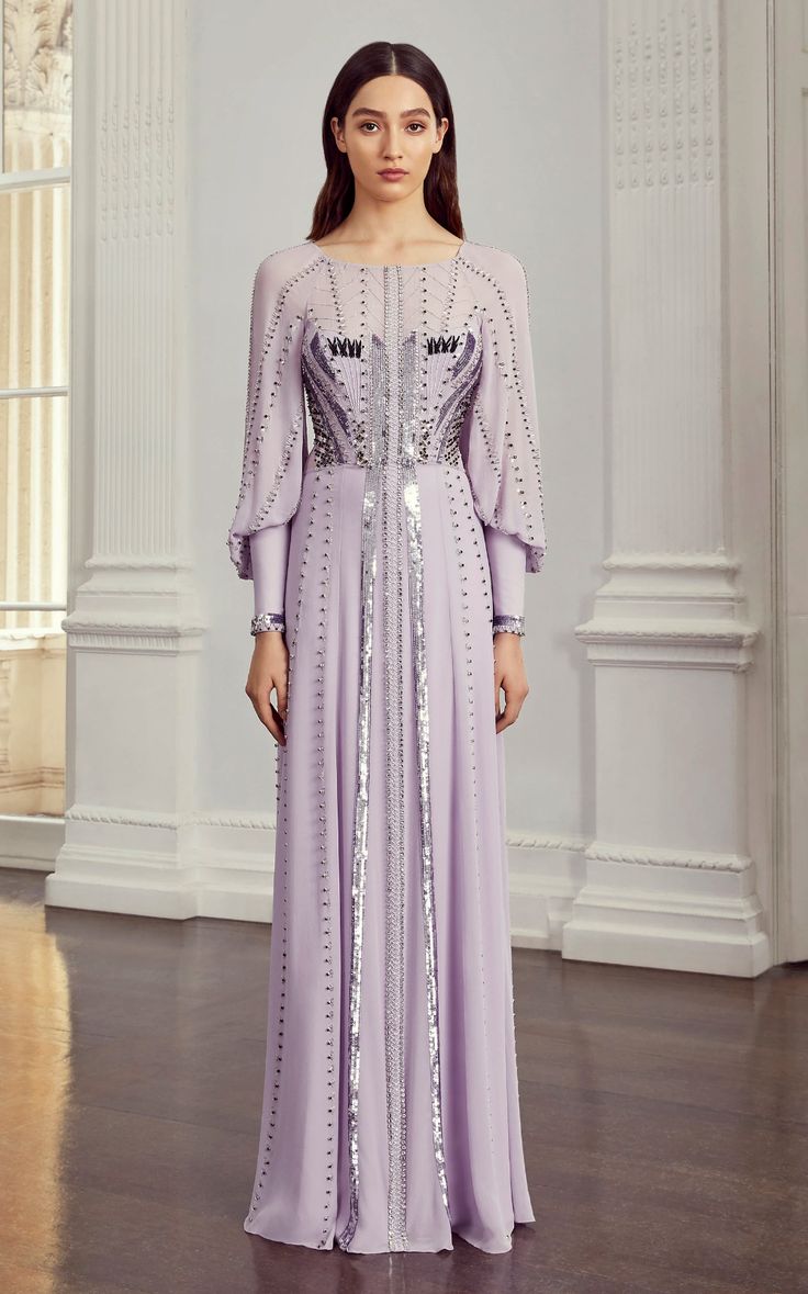 a fulllength studio shot of a female model wearing a stunning lavender maxi dress. the dress features long sleeves with a slight puff at the shoulder, a round neckline, and intricate sequin and bead embellishments. the embellishments form a vertical pattern down the bodice and sides of the dress, adding texture and visual interest. the model stands elegantly in a room with a classic white backdrop, adding to the luxurious feel of the image. the overall style is formal and sophisticated, suitable for special occasions such as weddings or galas. the dress is predominantly lavender or lilac in color, a soft and romantic hue. the fabric appears to be flowing and light, possibly chiffon or silk.