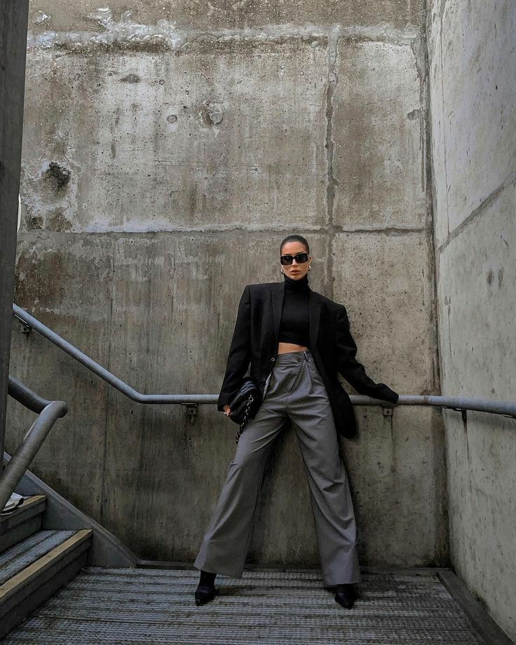 the image showcases a fashion model posing on a concrete staircase. she is wearing a stylish monochrome outfit consisting of a black oversized blazer, a black turtleneck, highwaisted wideleg grey pants, and black pointedtoe shoes. she is accessorized with black sunglasses and a small black handbag.  the background is a stark, urban setting of textured concrete walls and a metal railing. the overall aesthetic is minimalist and chic.