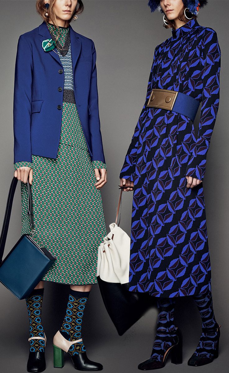 two female models are showcasing different outfits in a studio photoshoot. the model on the left is wearing a cobalt blue blazer over a green geometric print midi skirt. she's paired the look with patterned socks and unique twotoned heels. she carries a teal rectangular handbag and a creamcolored tote bag. the model on the right is wearing a longsleeved royal blue dress with a bold geometric pattern. the dress is cinched with a wide gold belt.  her look is completed with dark patterned socks and ankle boots. both models are styled with unique earrings and have stylish hair. the overall aesthetic is sophisticated and fashionable.