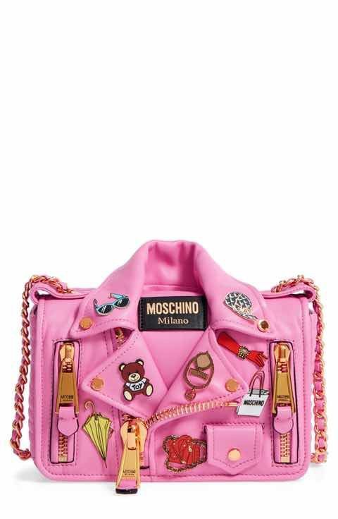 a pink leather moschino crossbody bag shaped like a biker jacket. it features a quilted leather body, metal studs and pins in the shape of a teddy bear, sunglasses, a moschino logo and other decorative accents. the bag has a chain strap and a flap closure.