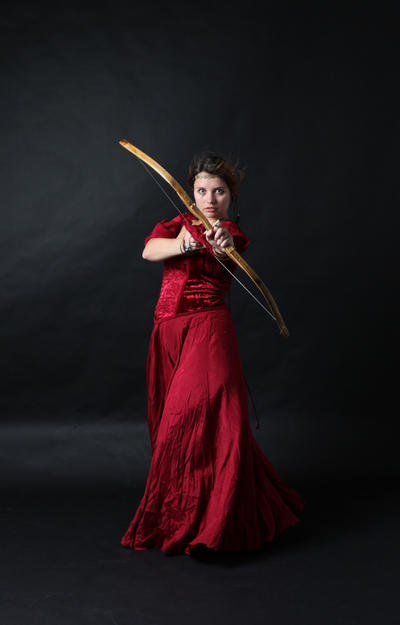a woman in a flowing red dress is captured in a dramatic studio shot. she is holding a bow and arrow, showcasing her archery skills with a poised and elegant stance. the dark background emphasizes the woman's red attire and the focus on her aiming position. the overall aesthetic is a blend of medieval fantasy and historical drama, suggesting a character from a tale or a theatrical production.