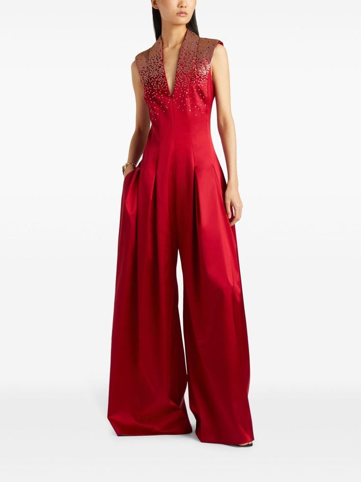 a woman is wearing a red sleeveless jumpsuit with a sequined collar. the jumpsuit has a vneck and wideleg pants. she is standing in front of a white background. she is looking at the camera and her hair is pulled back.