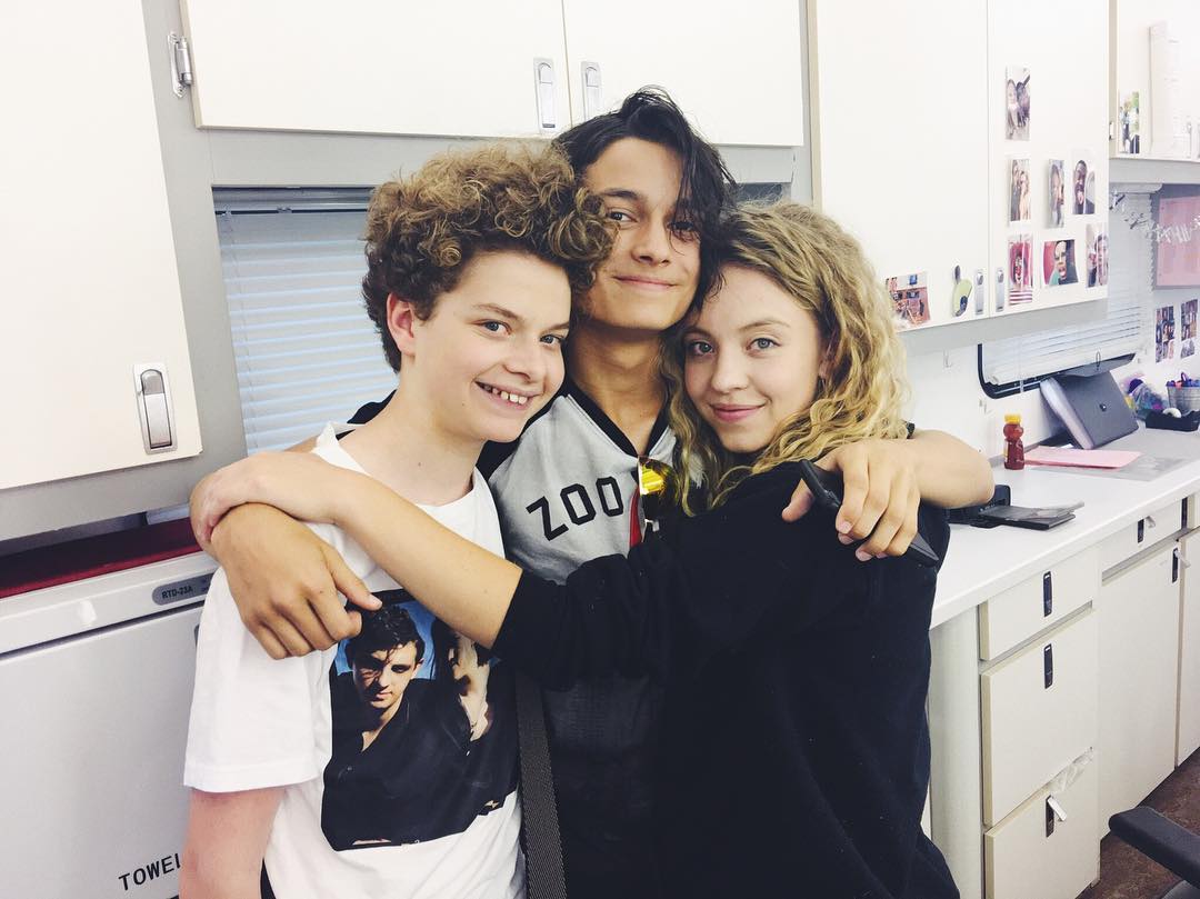 the image shows three teenagers embracing each other in what appears to be a film or television production's behindthescenes setting.  the background includes a kitchen area with cabinets and counters, indicating a break room or mobile unit.  one teenager in the center is wearing a shirt with a "zoo" logo. another teenager on the left has curly hair and wears a white tshirt with a picture of a man on it. the person on the right has long curly hair and is wearing a black top. all three are smiling and appear to have a friendly and close relationship. the overall atmosphere is casual and joyful.