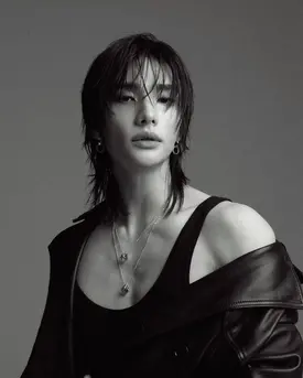 the image is a black and white fashion portrait of a male model with androgynous features. he has shoulderlength dark hair with bangs partially covering his forehead.  he is wearing a black sleeveless top and a black leather jacket draped over his shoulders.  he also wears several necklaces and small earrings. his expression is serious and intense. the background is a plain gray, creating a focus on the model. the overall aesthetic is edgy and stylish. the image has a highfashion, editorial feel.