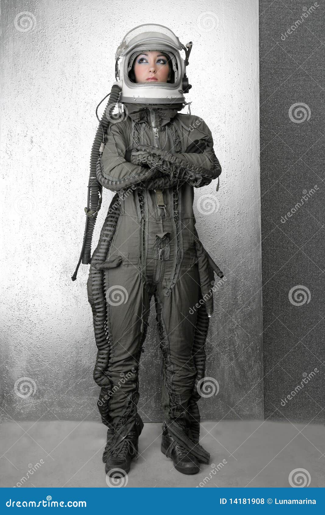 a woman dressed in an astronaut suit and helmet stands against a silver wall. she looks up with her arms crossed. her suit is black and gray with buckles and straps. she has a futuristic look and is surrounded by a scifi atmosphere.