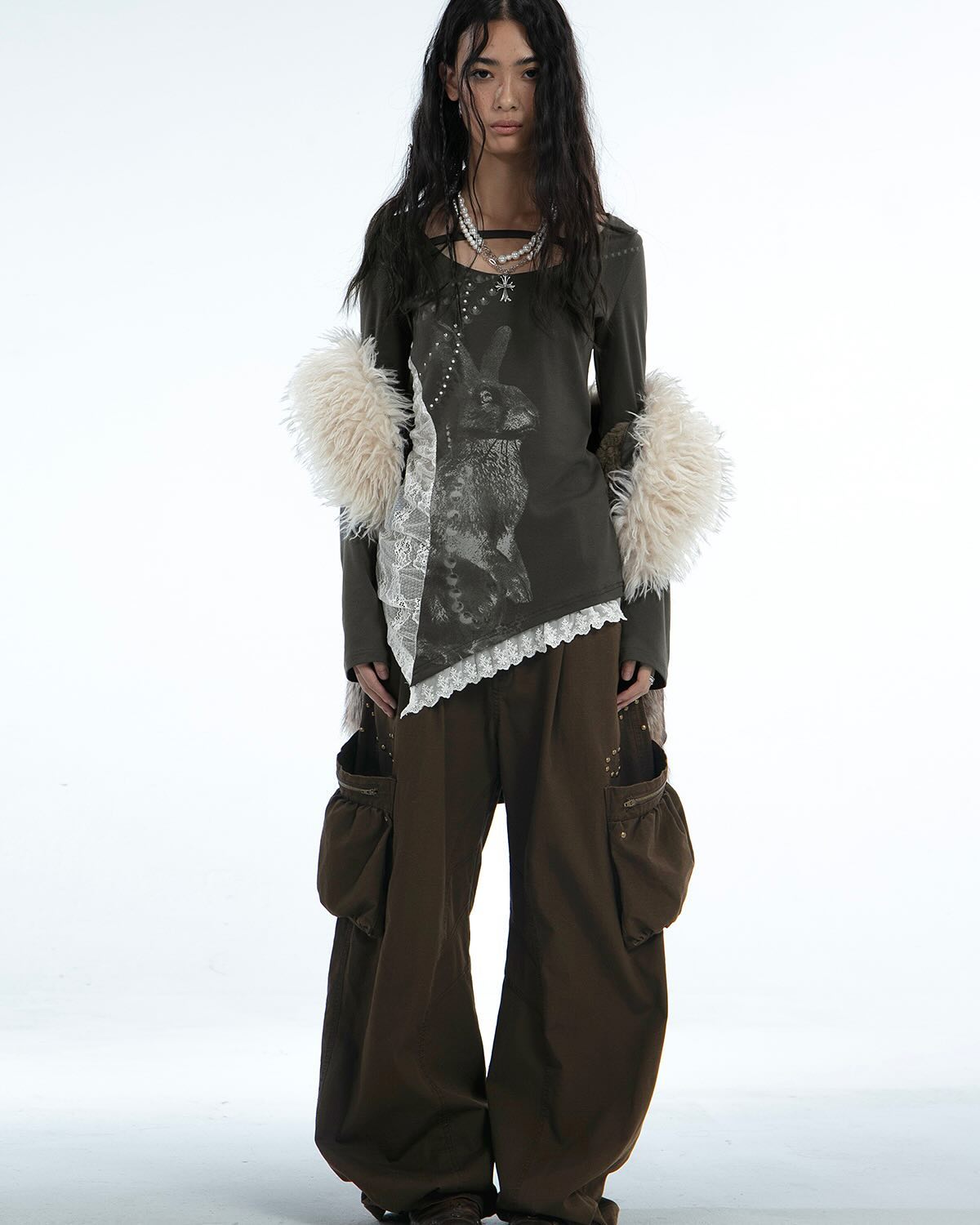 a fulllength studio shot of a female model wearing a dark olive green longsleeved top featuring a rabbit graphic print and lace detailing. the top is layered with a white faux fur trim around the shoulders. she is also wearing widelegged, dark brown cargo pants with added pocket details. the model has long dark hair and is wearing a delicate necklace. the background is plain white, focusing attention on the outfit.