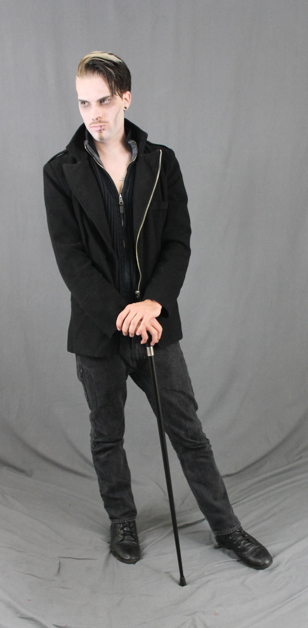a fullbody studio shot of a man with pale makeup and dark hair styled with blonde highlights. he is wearing a black coat, dark jeans, and black shoes. he holds a black cane. he's standing against a grey backdrop, and his expression is serious. the overall style is gothic or alternative.