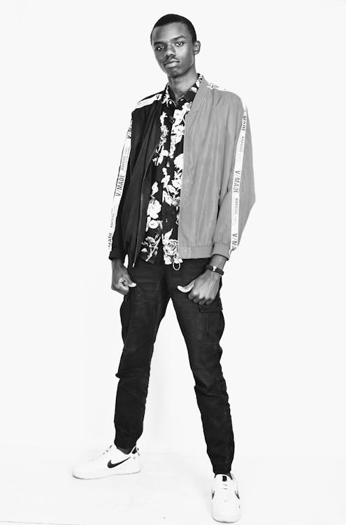 a black and white fullbody studio shot of a young man. he is standing and looking at the camera. he is wearing a floral shirt under a black and light gray bomber jacket with white stripes on the sleeves. he has on black cargo pants and white sneakers. the background is white.