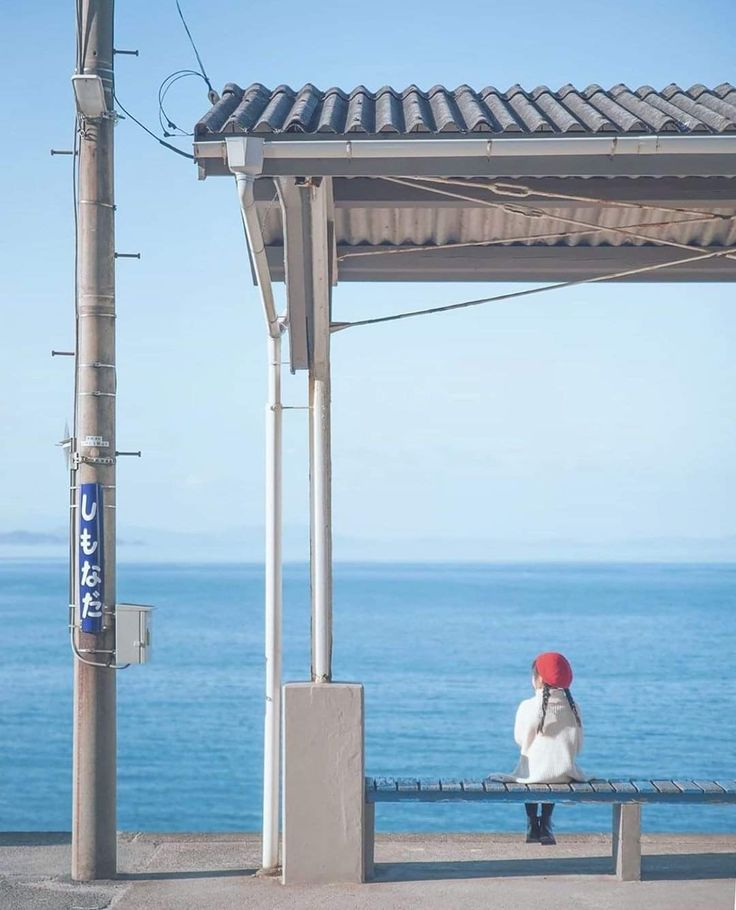 a girl wearing a red beret and white coat sits on a blue bench under a roofed bus stop, looking out at a calm blue sea and sky. a utility pole stands beside the bus stop. the sign on the pole reads "shimada". the horizon line is visible in the distance.