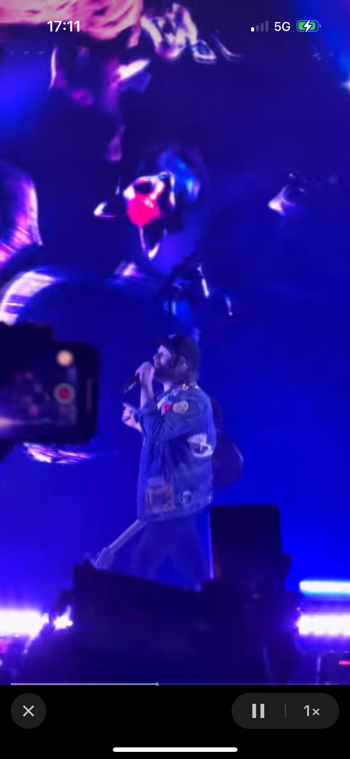 a concert singer is performing on stage, he is holding a microphone and singing into it. he is lit up with blue spotlights, with the rest of the stage dark. the background is blurry with what looks like a crowd in the background.