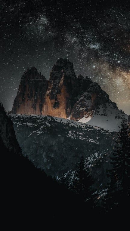 the image showcases a dramatic nighttime landscape featuring a towering mountain range under a starry sky. the mountains, some capped with snow, are rugged and imposing. the sky is filled with countless stars, and the milky way stretches across the horizon, creating a breathtaking cosmic view. a dark forest with fir trees is visible in the foreground, adding depth and framing the majestic mountain peaks. the overall atmosphere is one of awe and wonder, capturing the beauty and grandeur of nature at night.
