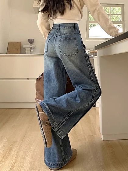 a woman is shown from the back, wearing wideleg, highwaisted denim jeans. the jeans have a light wash and button details on the side. she is also wearing beige mules. the photo appears to be taken indoors, with a wooden floor visible.