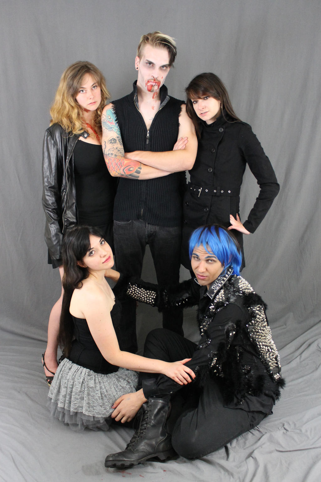a group of five people, three women and two men, pose for a group photo against a gray backdrop. their attire consists primarily of black clothing in various styles of goth and alternative fashion. the man in the center has vampire makeup. one woman wears a leather jacket, another a black dress, and a third wears a gray tutustyle skirt. the two men are dressed in black, one in studded attire, the other more casual. the overall impression is a band or group of friends showcasing their dark and alternative style.