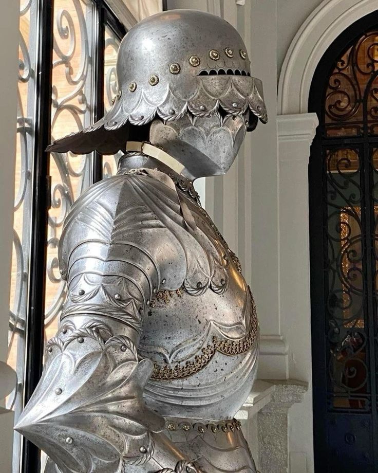 a detailed closeup shot of a suit of antique, ornate, silvergrey medieval armor. the armor is highly detailed with intricate engravings and embellishments, showcasing exceptional craftsmanship. the armor includes a helmet with a decorative brim and a body piece that covers the torso and arms, partially showing the pauldrons and gauntlets. the armor is displayed in an elegant interior setting, with a glimpse of a decorative door and window with wroughtiron detailing in the background, emphasizing the historical and luxurious context. the lighting enhances the metallic sheen and the threedimensional texture of the armor.