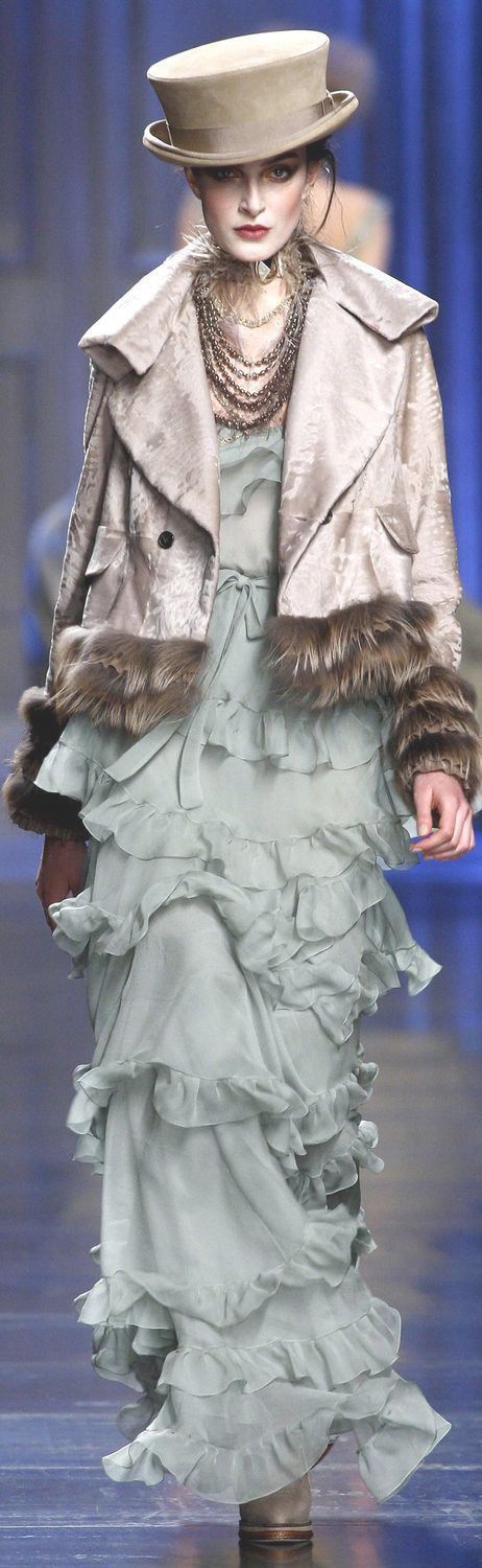 a runway model is shown wearing a light blue dress with layered ruffles and a taupe jacket with fur trim. she also has on a matching hat and numerous necklaces. the background is a blue stage.