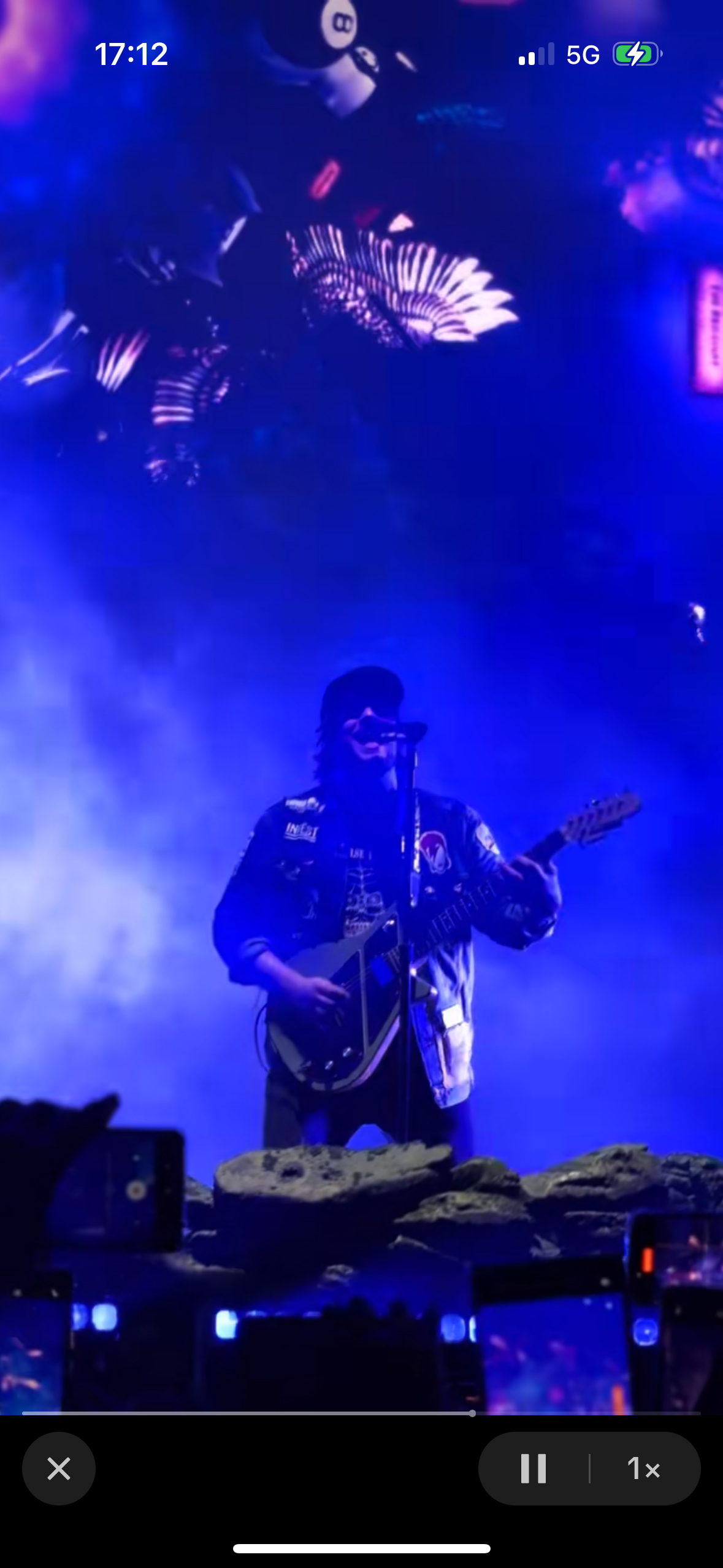 a musician on stage playing a guitar under a spotlight, facing the audience.  the image is blurry due to motion. the person is wearing a dark jacket and jeans, and you can see a microphone on their side. they are performing in front of a large crowd, but the crowd is blurred, and the stage is dark.  the lights behind the performer are blue and purple, creating a dreamy atmosphere.  the musician looks happy and excited to be performing, and the crowd is clearly enjoying the show.