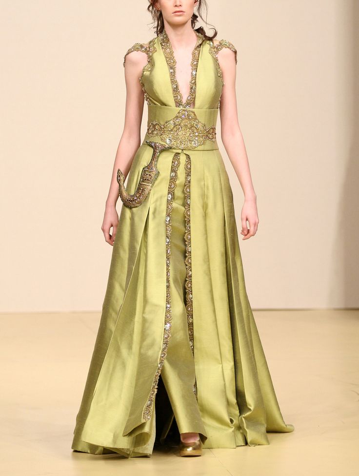 a full shot of a female model showcasing a stunning lime green gown on a runway. the dress features intricate gold embroidery and embellishments, including a jeweled belt and a decorative dagger at the waist.  the gown is a long, flowing design with a high slit, a vneckline, and sleeveless cut. the model's pose and the setting suggest a highfashion, possibly couture, presentation. the overall aesthetic is elegant and sophisticated.