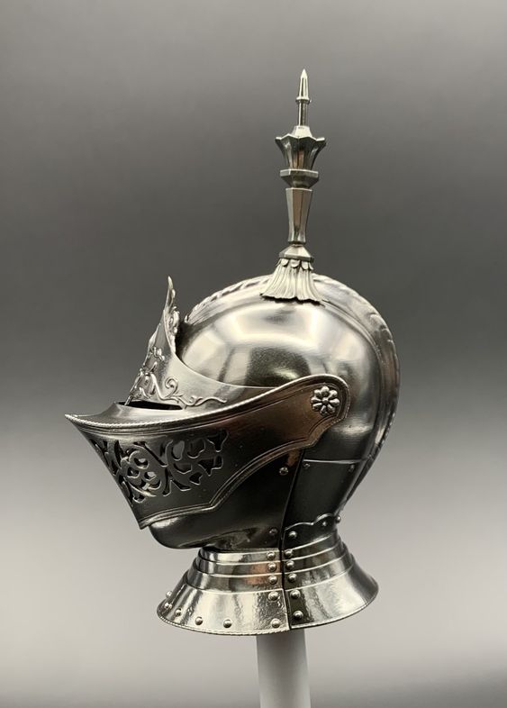 this is a closeup image of a knight's helmet in silver color, set against a light gray background. the helmet is intricately designed with floral patterns on the visor and neck guard, and a tall, pointed finial on top. the lighting highlights the metallic sheen and the detailed craftsmanship of the piece.