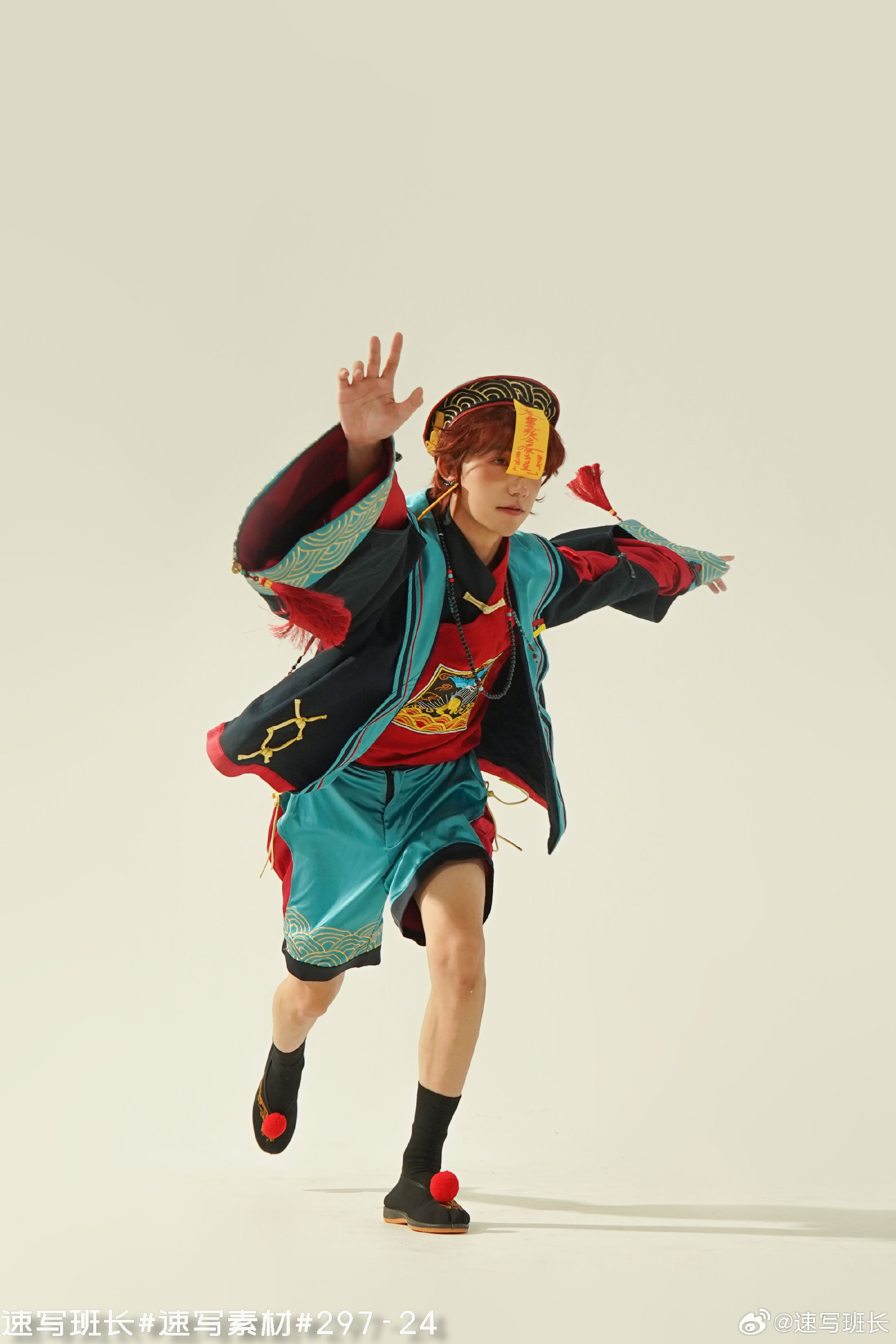 a man in an elaborate, colorful costume is captured midpose, as if dancing or performing a martial arts move, against a plain white background in a studio setting. the costume includes a black jacket with colorful accents, a vibrant teal undergarment, and a red headband with yellow tassels. the pose suggests movement and energy. the artificial lighting illuminates the scene, highlighting the man's dynamic posture.