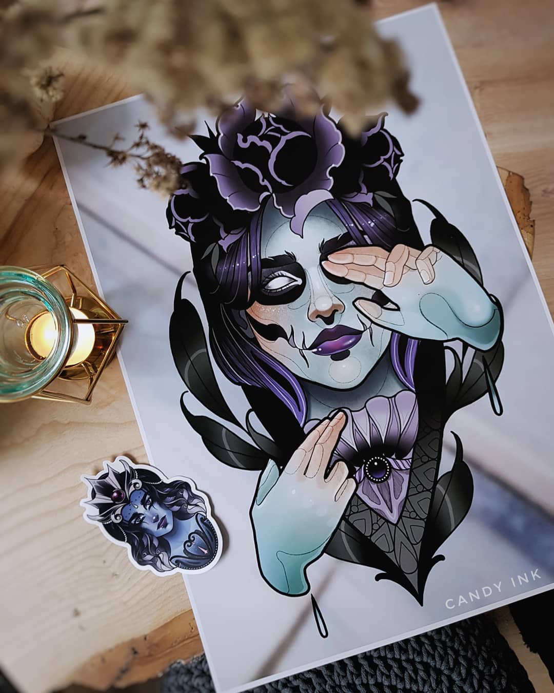 a print of a gothic illustration of a woman with purple hair, pale blue skin, and dark makeup wearing a crown of black flowers. she is holding her hands up to her face. there is a smaller sticker of the same woman on the paper as well. both are on a table with a lit candle and a knit object.