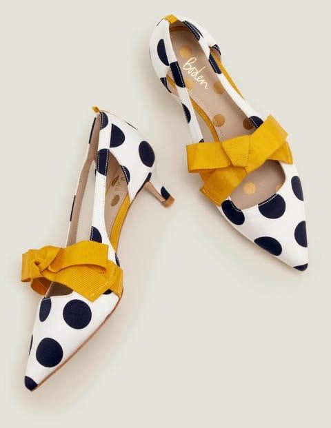 the image showcases a pair of boden brand women's shoes. they are white with navy blue polka dots and a bright yellow bow on each shoe. the shoes are high heels.
