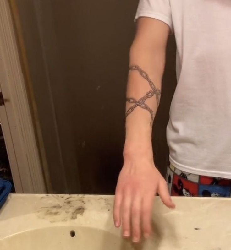 a man with a chain tattoo on his arm stands in a bathroom with a white shirt on, he is in front of the sink with his hand hovering over the sink.