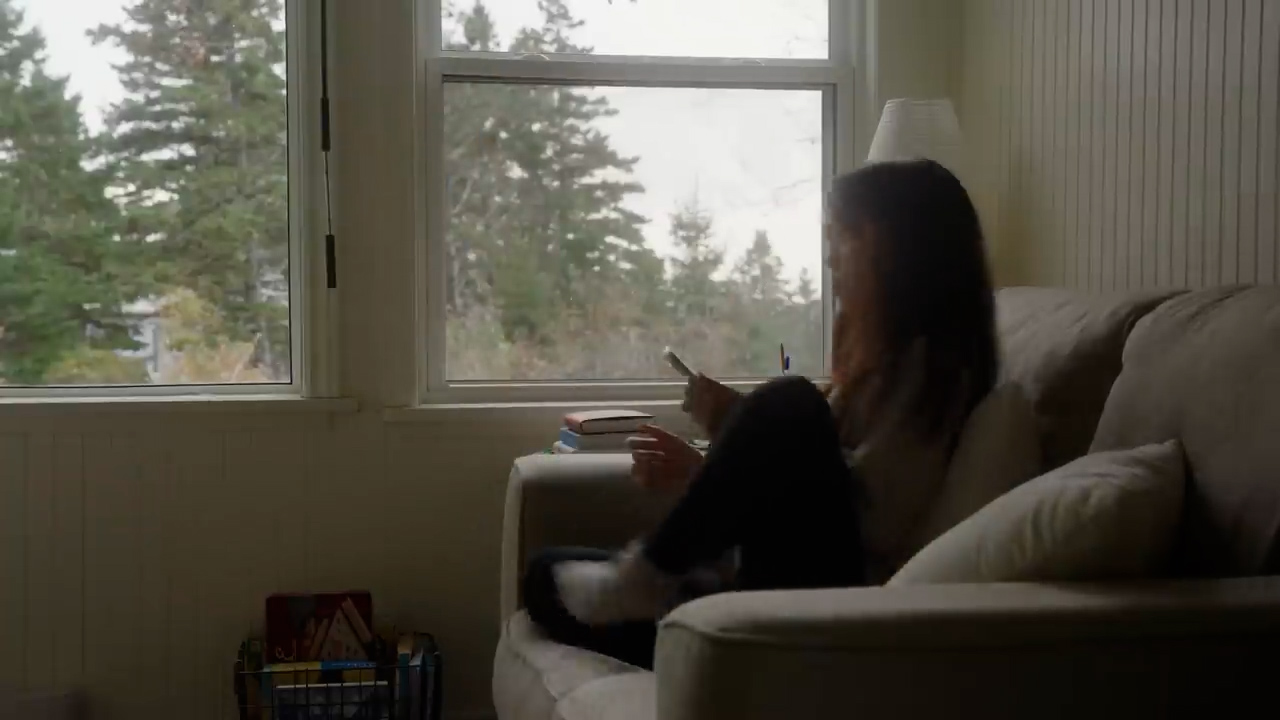 a young woman is curled up on a lightcolored couch in a living room, bathed in soft, natural light from two large windows. she is looking at her phone while books rest on the arm of the couch beside her. the windows offer a glimpse of a serene view of evergreen trees, suggesting a quiet and peaceful atmosphere.
