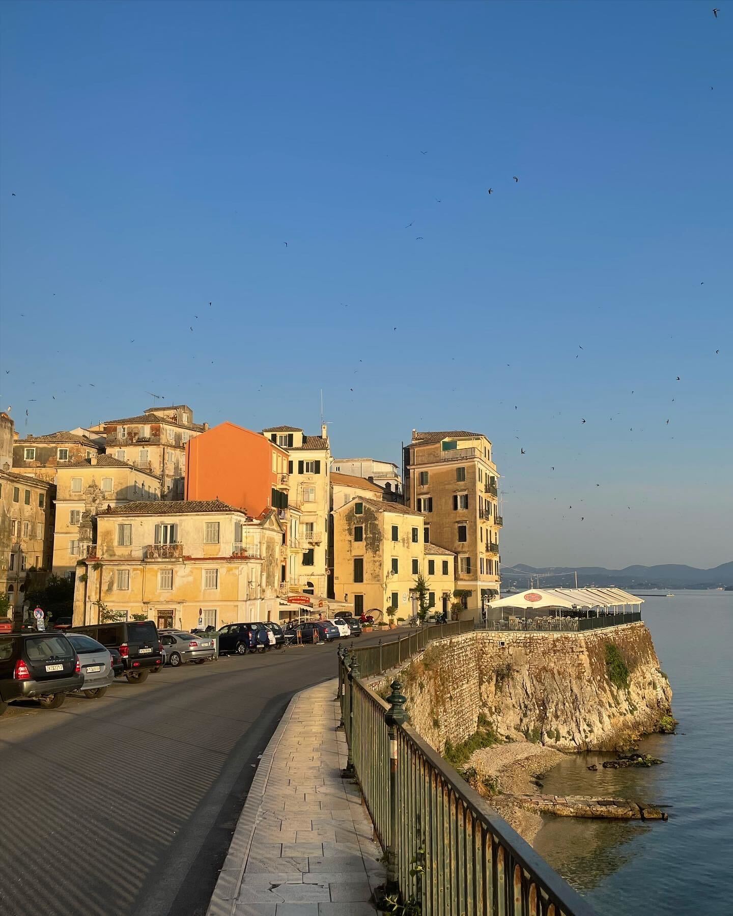 a picturesque view of a greek town on the waterfront. buildings line the street that slopes down to the sea. the street is lined with parked cars and the sea is dotted with boats. the town is bathed in the warm light of the setting sun and the blue sky is clear and cloudless. the image captures the beauty and charm of corfu town, greece.