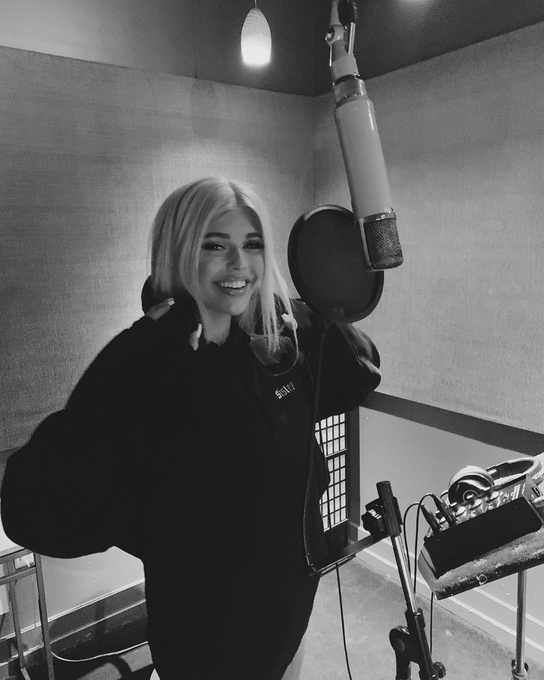 a black and white image of a woman smiling in a recording studio standing next to a microphone with a pop filter. there are headphones and other audio equipment in the background. the woman has blonde hair and is wearing a dark colored hooded sweatshirt.
