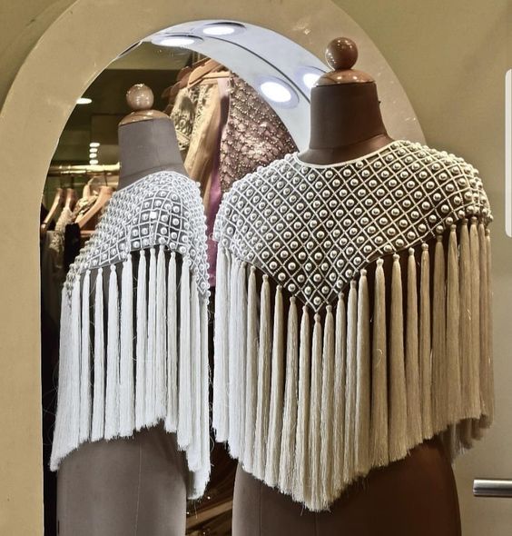 two mannequins stand sidebyside, displaying elegant white capes. the capes feature intricate beadwork, likely pearls, arranged in a grid pattern across the upper portion. long, flowing tassels of white fringe cascade from the bottom edge, creating a dramatic and sophisticated silhouette. the mannequins are positioned within a clothing store, suggesting a retail setting.